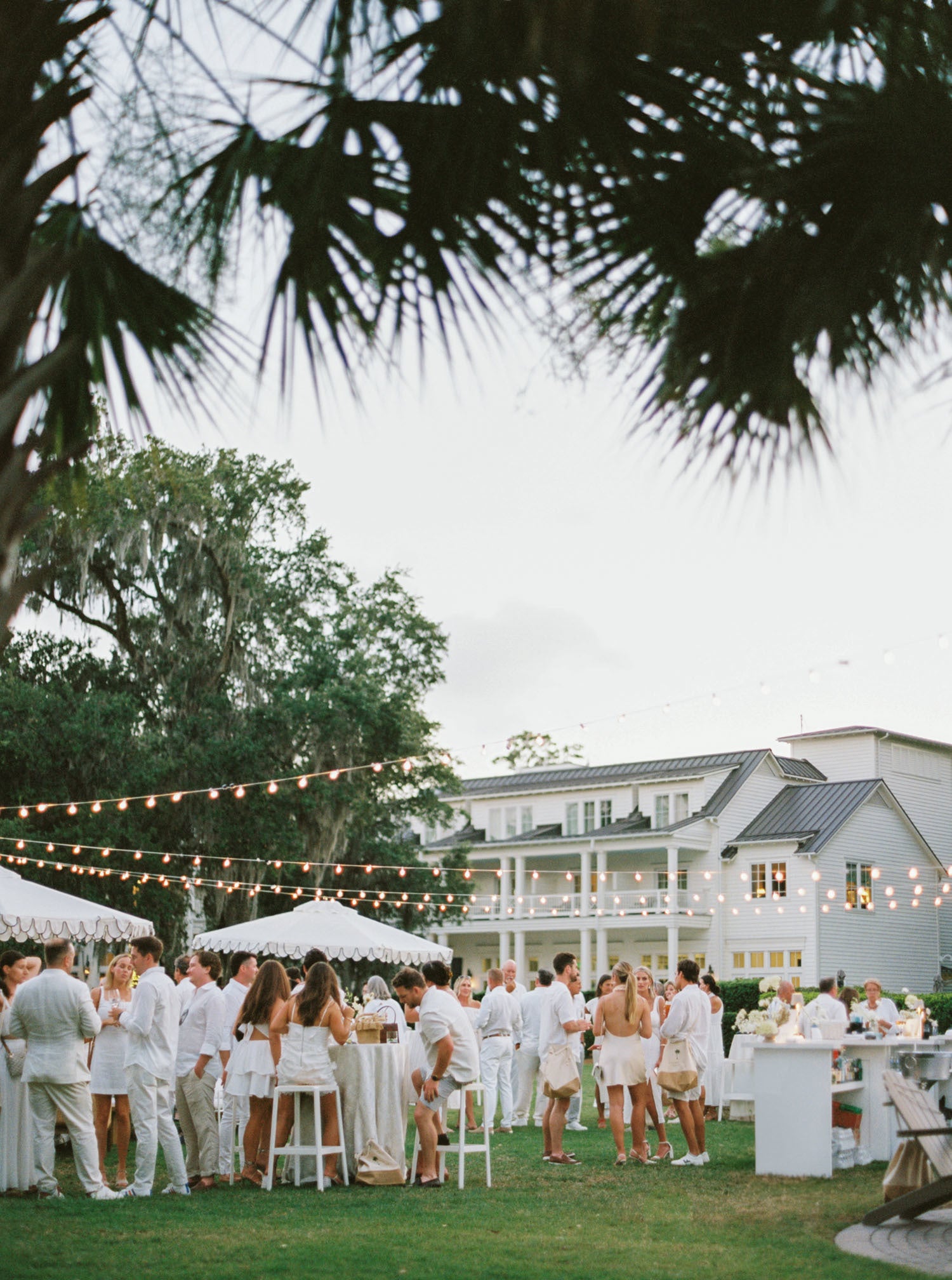 Outdoor wedding reception with guests in white attire, string lights, and a large building in the background.