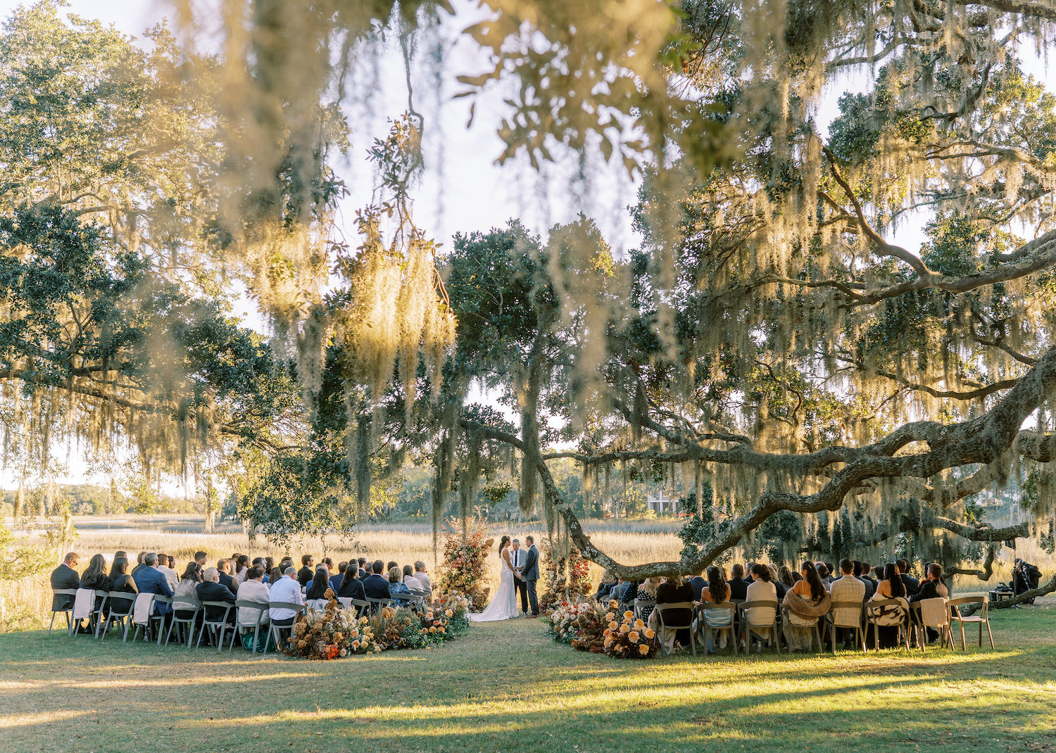 Outdoor wedding ceremony beneath oak trees at Kiawah River in South Carolina, with neutral-toned guest seating by Mayker Events and organic floral aisle installations by Blossom Bay Design.