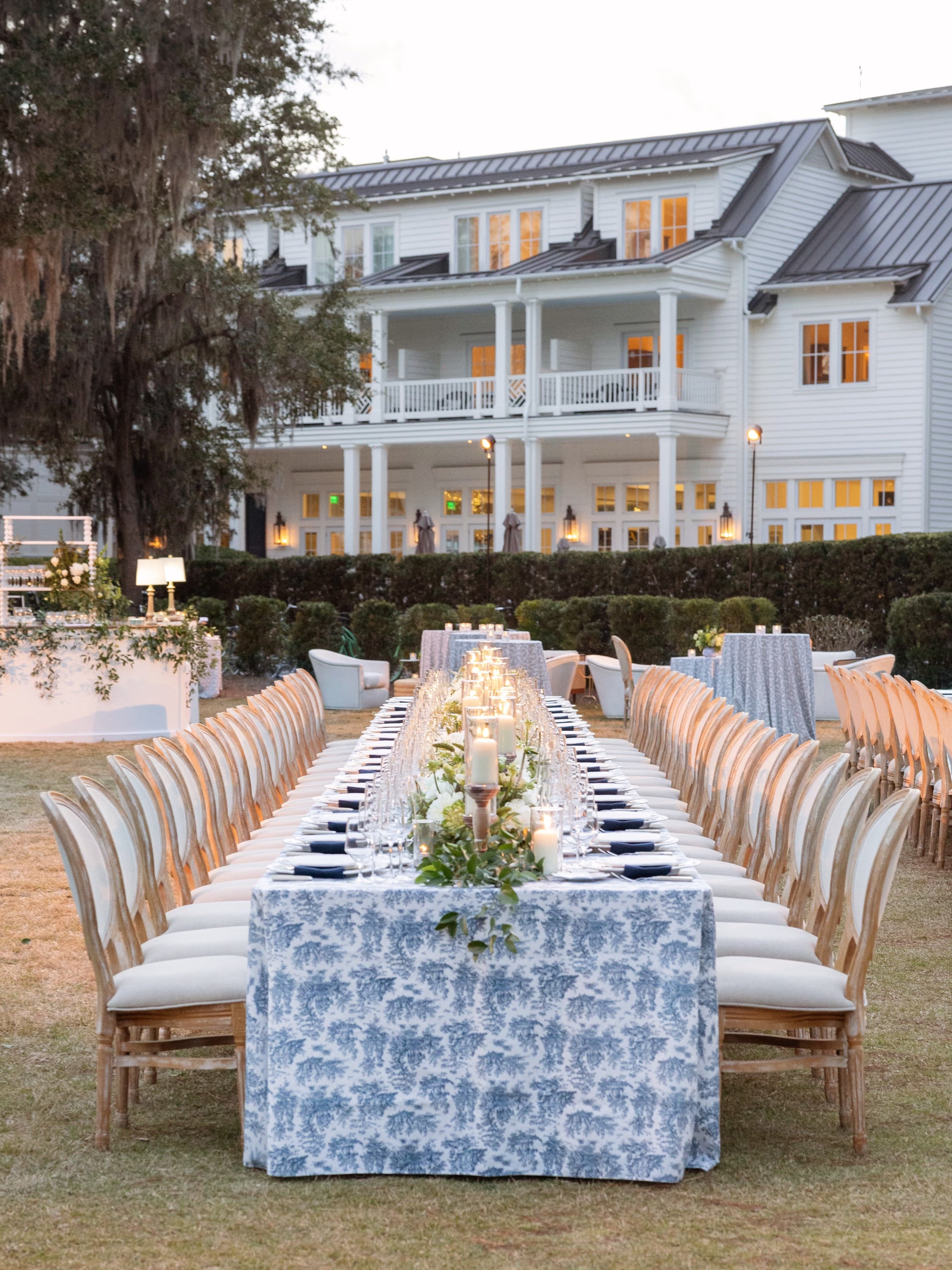 Elegant outdoor dinner reception designed by Meredith Tanton Events, with long blue toile-covered tables, classic chairs, and soft candlelight in front of a white Southern estate.