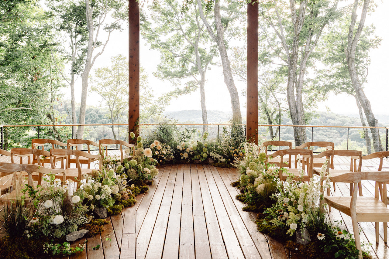 Outdoor wedding setup at Southall Farm,  designed by Kelsey Rae Designs, with wooden chairs, tables, and floral arrangements on a wooden deck with trees in the background.