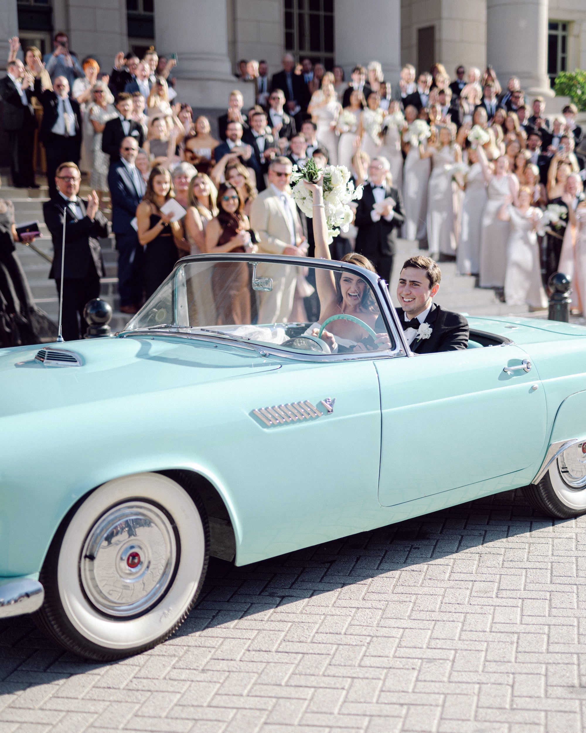 Light blue vintage car with newlyweds inside, surrounded by people in formal attire on a brick pavement. Shot by Kéra Golumbisky.